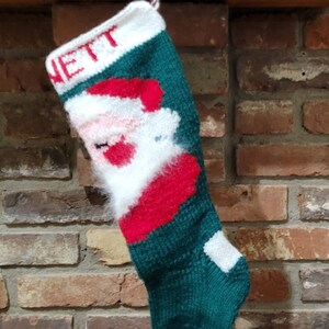May include: A green, red, and white knitted Christmas stocking with a Santa Claus design and the name "NETT" embroidered on the top.
