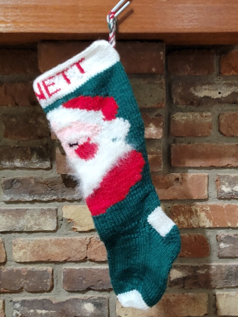 May include: A green, red, and white knitted Christmas stocking with a Santa Claus design and the name "NETT" embroidered on the top.