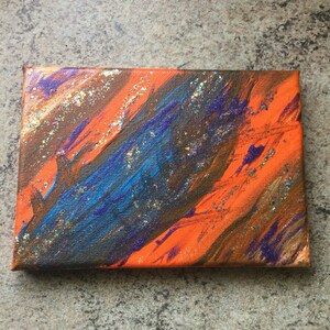 May include: Abstract painting on canvas with orange, blue, brown, and purple paint. The painting has a textured surface with glitter embedded in the paint.