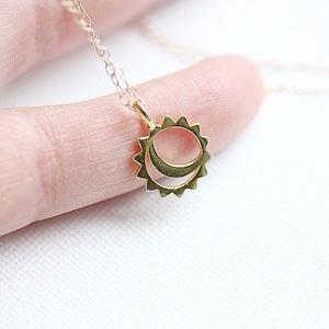 May include: A gold-coloured necklace with a pendant depicting a sun and crescent moon. The pendant is small and detailed, with a textured surface. The necklace is shown against a soft, neutral background.