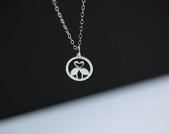 May include: A silver-toned necklace featuring a circular pendant with a cutout design of two flamingos facing each other, their necks forming a heart shape. The pendant hangs from a delicate silver-toned chain with small, uniform oval links.