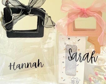 May include: Two clear plastic tote bags with handles. The bag on the left has a black bow and the name "Hannah" written on it. The bag on the right has a pink bow and the name "Sarah" written on it.