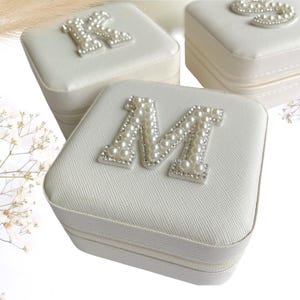 May include: White square jewelry box with a zipper closure. The box has a pearl-embellished letter "M" on the top.