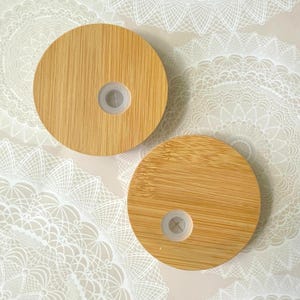 May include: Two round bamboo lids with a light wood grain pattern. Each lid has a central, clear plastic insert. The lids are designed for a drinking glass or bottle. The background is a light beige color with a white lace pattern.