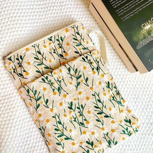 May include: Cream-coloured daisy embroidered pouch with a matching strap and zip closure. The pouch is next to a book with the title "Chaos Walking". The daisies have white petals and yellow centres, with green stems and leaves.