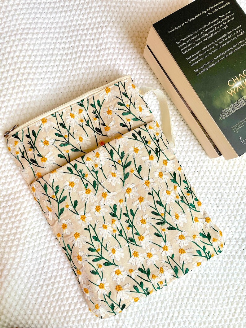 May include: Cream-coloured daisy embroidered pouch with a matching strap and zip closure. The pouch is next to a book with the title "Chaos Walking". The daisies have white petals and yellow centres, with green stems and leaves.