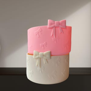 May include: A two-tiered cake-shaped container. The top tier is pink, and the bottom tier is off-white. Both tiers feature embossed bow patterns. A large pink bow adorns the top, and a white bow is on the bottom tier.
