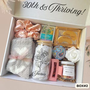May include: A gift box labeled 30th & Thriving containing a peach satin scrunchie, gray fuzzy socks, a glass tumbler with a floral design and the name Jamie, a tea bag, gold eye masks, a paisley tin, a white rose, a pink hair clip, and bath salts.
