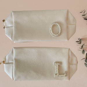 May include: Two white cosmetic bags with pearl-embellished letter charms. The top bag has a pearl "O" charm and the bottom bag has a pearl "L" charm.