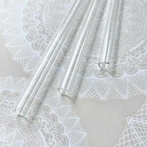 May include: Three clear glass drinking straws on a white lace background.
