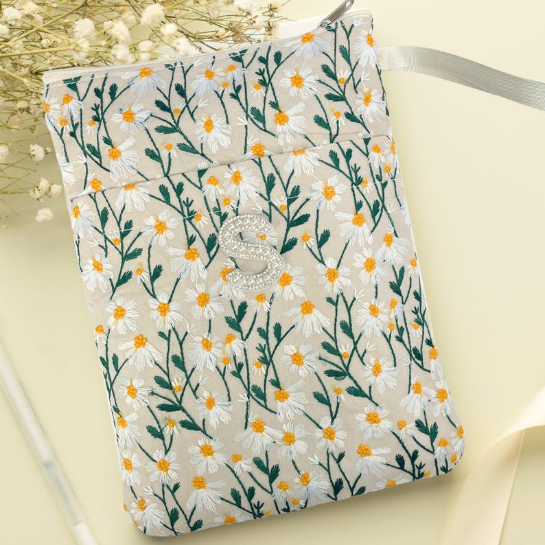 May include: A beige embroidered pouch with a daisy floral pattern. The pouch features white daisies with yellow centres and green stems. It has a silver zip and a rhinestone-studded silver initial "S". A light grey ribbon is attached.