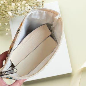 May include: A beige floral print book sleeve with a silver zip, containing two books. The interior is white. The sleeve features yellow and green floral patterns. The background includes a white book and a light yellow surface.