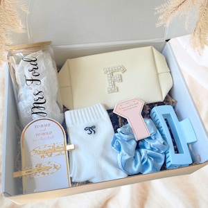 May include: A gift box filled with various items. Includes a cream-colored clutch with a jeweled "F", a glass jar with "Mrs Ford" written on it, white socks with a bow, a light blue hair claw clip, and a light blue scrunchie.