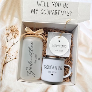 May include: A gift box with a frosted glass tumbler, a white mug, and a round ornament. The tumbler is labeled "Godmother Est. 2026", the mug says "Godfather Est. 2026", and the ornament reads "Godparents Est. 2026". The box lid reads "Will You Be My Godparents?"