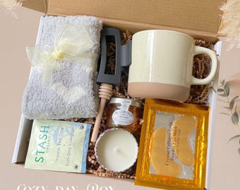 May include: A gift box filled with self-care items. Includes a gray plush sock with a yellow bow, a cream-colored mug with a tan base, a jar of honey, a candle, tea, and eye masks. The box is labeled "COZY DAY BOX".