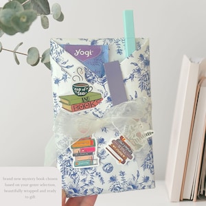 May include: A gift-wrapped package with a blue and white floral pattern, tied with a white ribbon. The package contains a tea bag, a bookmark, and a pen. Stickers with the words "cup of tea and books" and "books is still a romance" are visible.