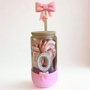 May include: A clear glass jar with a wooden lid, filled with pink fabric and a decorative silver-rimmed oval. A pink bow adorns the bottom and top. A pink bow-topped straw extends from the lid.