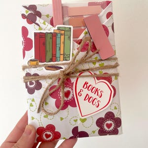 May include: A wrapped gift with a floral pattern in white, red, and purple, tied with twine. The gift is adorned with stickers of books, a coffee cup, and a heart-shaped tag that reads "BOOKS & DOGS". Pink bookmarks and sticky notes are visible.