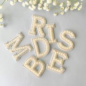 May include: Pearl-covered letters spelling "BRIDE" on a light blue surface, with a scattering of white flowers in the background. Each letter is outlined with silver rhinestones. The letters are in a serif font.