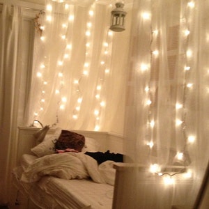 May include: A white bed with a white headboard and a white duvet cover. The bed is decorated with white sheer curtains and string lights. The lights are strung across the top of the bed and create a cozy and romantic atmosphere.
