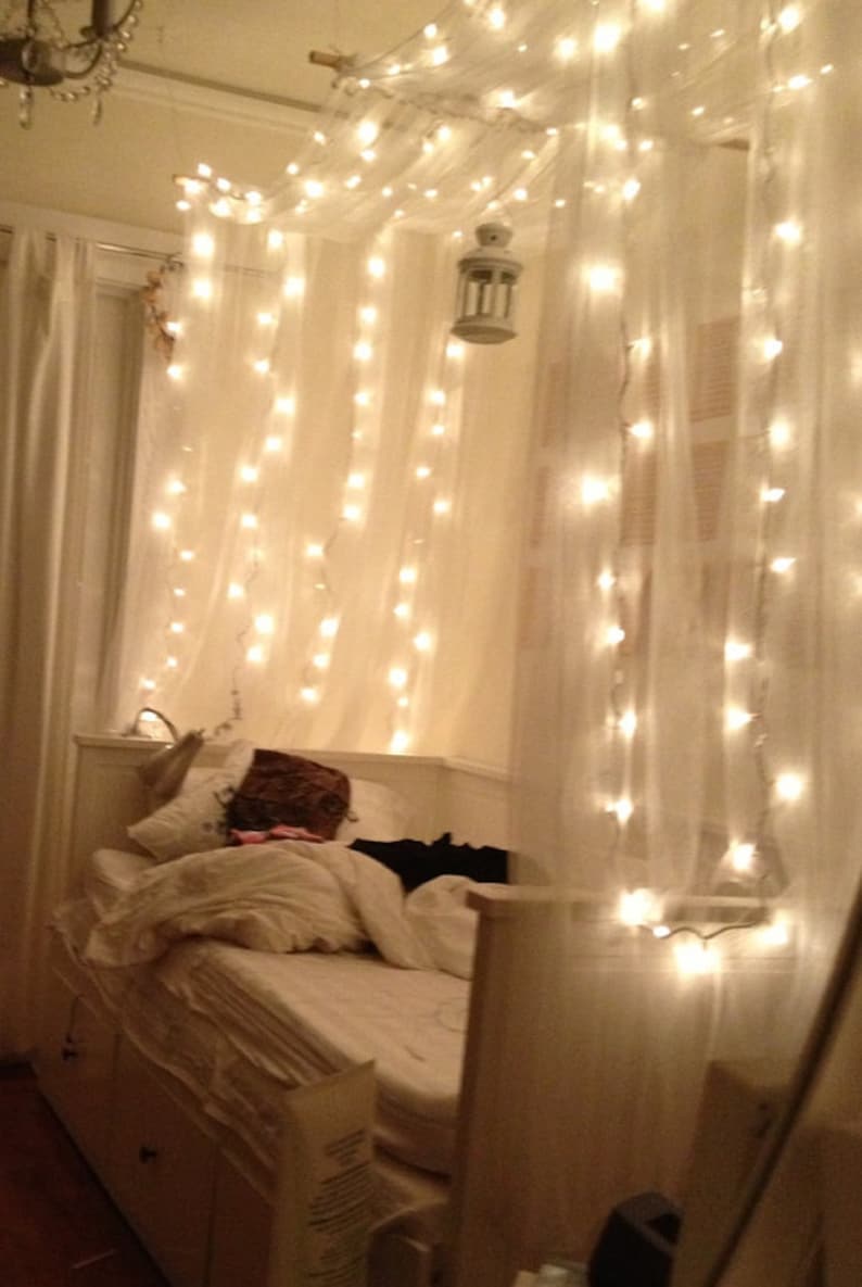 May include: A white bed with a white headboard and a white duvet cover. The bed is decorated with white sheer curtains and string lights. The lights are strung across the top of the bed and create a cozy and romantic atmosphere.