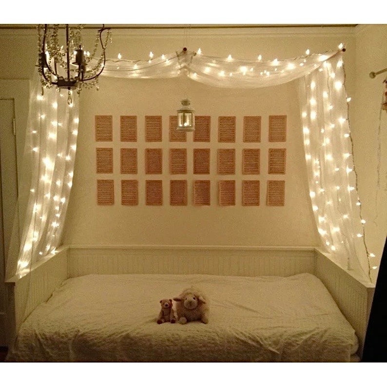 May include: A white canopy bed with white string lights draped over it. The bed has a white headboard and footboard and a white mattress. There are two stuffed animals on the bed, a white sheep and a brown pig. The bed is in a room with a white wall and a wooden ceiling.