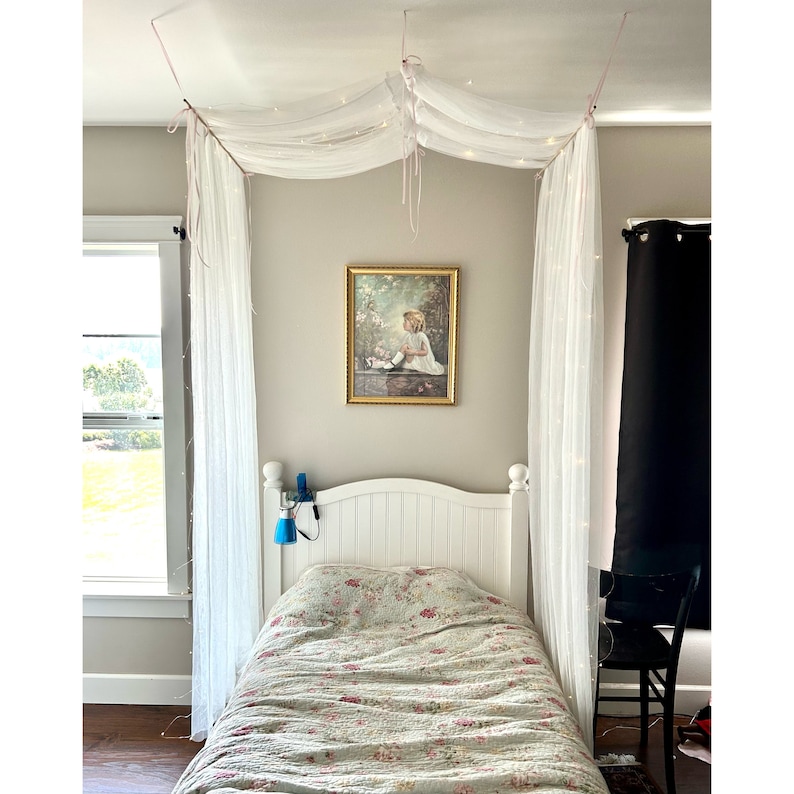 May include: A white bed with a floral patterned comforter and a white canopy with fairy lights. A framed picture of a girl in a white dress is hanging on the wall behind the bed.