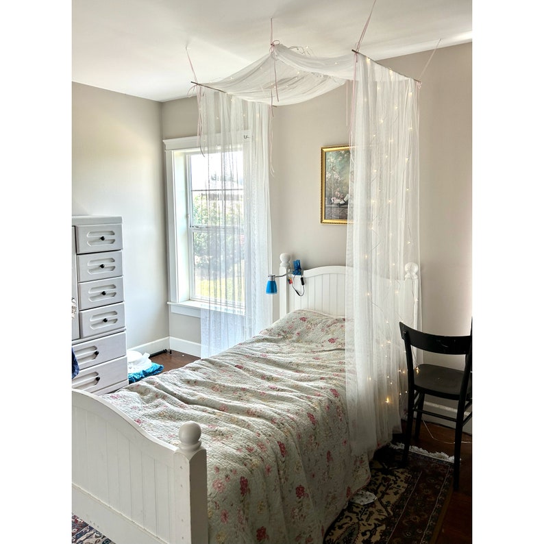 May include: A white bed with a white canopy and string lights. The bed has a floral patterned comforter and a white headboard. There is a dresser and a chair in the room.