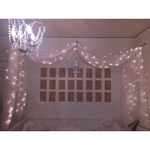 May include: A white canopy bed with sheer white curtains and string lights. A white lantern hangs from the ceiling above the bed. The wall behind the bed is decorated with a grid of brown squares.