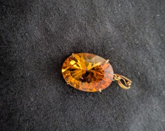 May include: A gold pendant featuring a large, oval-cut, amber-colored gemstone. The gemstone has multiple facets that reflect light, creating a sparkling effect. The pendant is set in a gold metal setting.