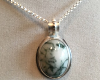 May include: A silver pendant necklace featuring an oval-shaped moss agate stone with a smooth, polished surface. The stone displays a creamy white base color with intricate, dark green, fern-like inclusions throughout. It hangs from a silver chain.