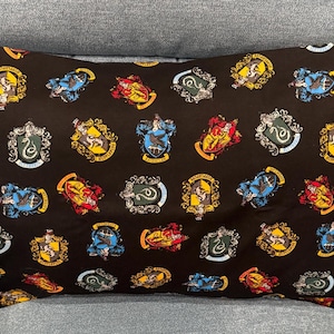 May include: A rectangular pillow with a dark brown background, covered in repeating crests. The crests feature various colors including red, gold, blue, and green, with intricate details and a decorative border. The pillow is resting on a gray surface.