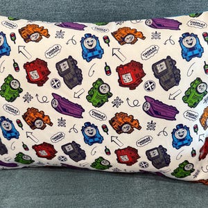 May include: A rectangular pillow with a white background featuring a repeating pattern of colorful cartoon trains, traffic lights, arrows, and the text "Thomas & Friends." The trains are in various colors, including blue, orange, green, red, and purple.