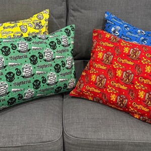 May include: Several decorative throw pillows with patterns representing different houses from a fantasy series. The pillows feature the house names and crests in yellow, green, red, and blue, resting on a gray couch.