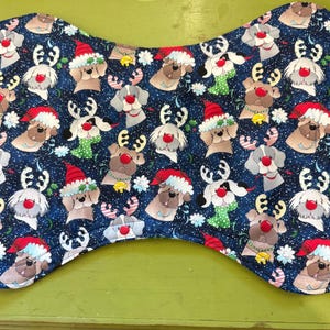 May include: Bone-shaped pet placemat with a festive pattern. The navy blue fabric is covered with illustrations of dogs wearing Santa hats, reindeer antlers, and red noses. White snowflakes and candy canes are scattered throughout the design.
