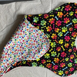May include: A reversible dog bandana with a black background and rainbow paw prints and bones on one side and a white background with multi-colored paw prints and bones on the other side. The bandana is folded in half and the tag is visible with the text "X-Large".
