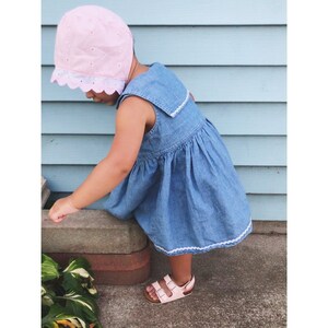 May include: A young child wearing a pink sun hat with white eyelet trim and a blue denim dress with white trim. The child is standing on a concrete surface and looking down.