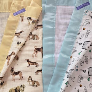 May include: Two baby blankets with satin borders. One has a yellow border and a dog pattern, the other has a light blue border and a sports-themed pattern. Both blankets have a tag that reads "A World 2 Day 2 Be".