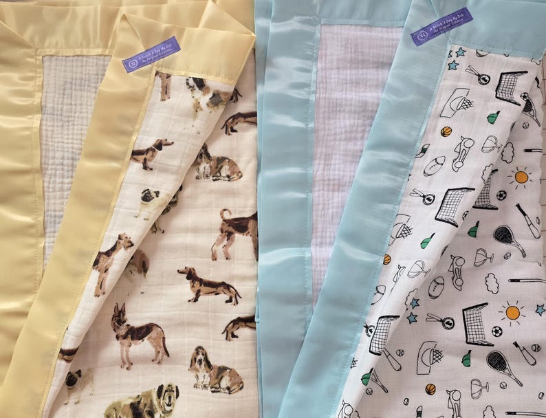 May include: Two baby blankets with satin borders. One has a yellow border and a dog pattern, the other has a light blue border and a sports-themed pattern. Both blankets have a tag that reads "A World 2 Day 2 Be".