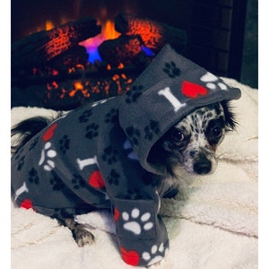 May include: A small dog wearing a gray fleece hooded sweater with paw prints and red hearts. The sweater has the text "I love" on it. The dog is sitting on a white fluffy surface.