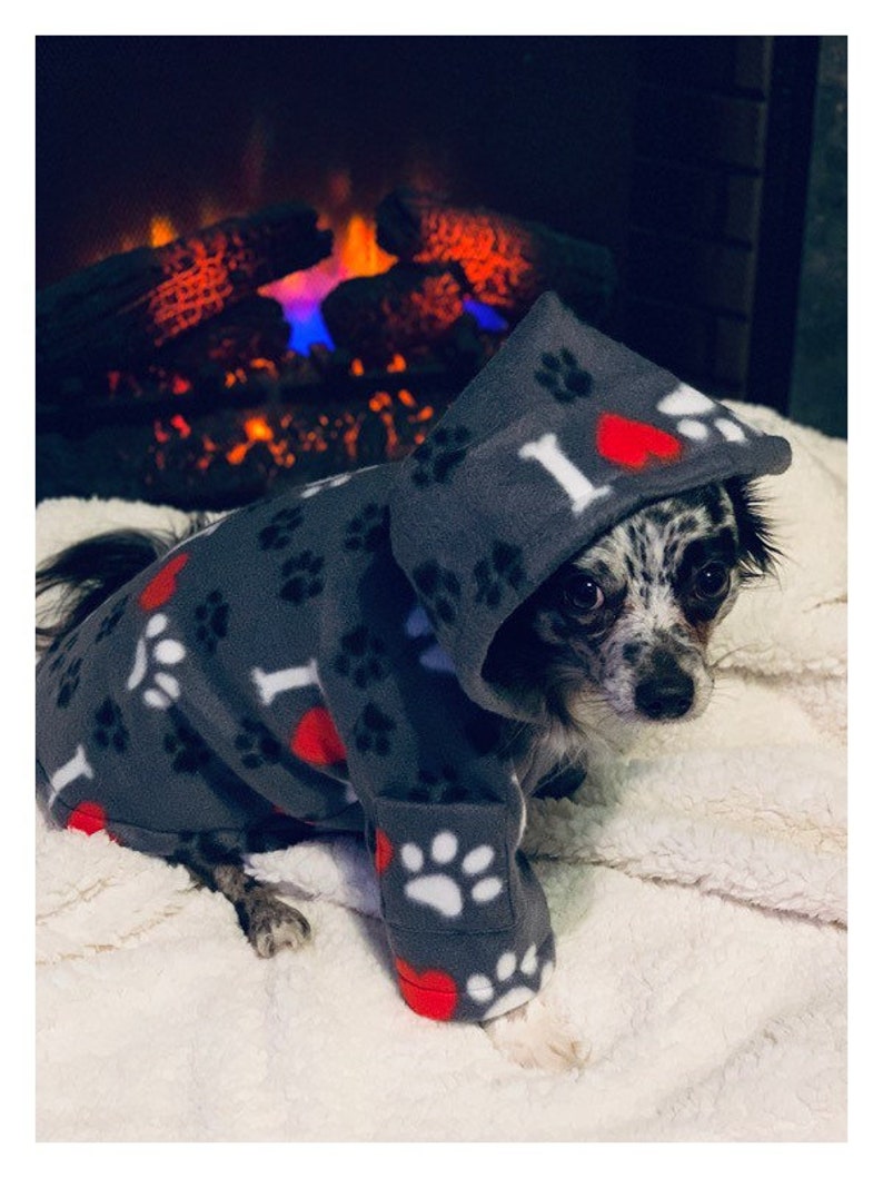 May include: A small dog wearing a gray fleece hooded sweater with paw prints and red hearts. The sweater has the text "I love" on it. The dog is sitting on a white fluffy surface.