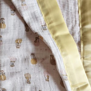 May include: A baby blanket with a cream-colored, patterned interior featuring hot air balloons in shades of gold, pink, and brown. The blanket is trimmed with a soft, yellow satin border. A blue tag with white text is sewn into the corner.
