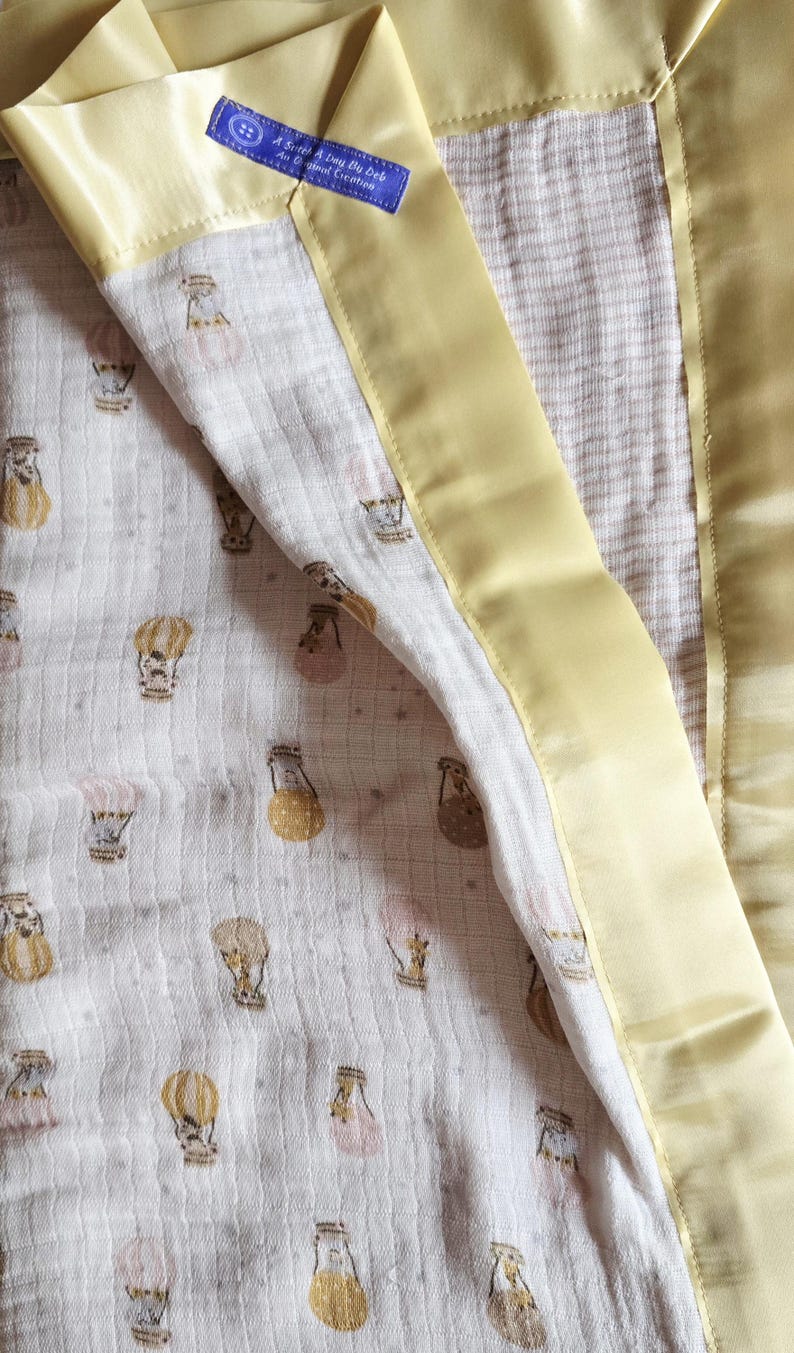 May include: A baby blanket with a cream-colored, patterned interior featuring hot air balloons in shades of gold, pink, and brown. The blanket is trimmed with a soft, yellow satin border. A blue tag with white text is sewn into the corner.