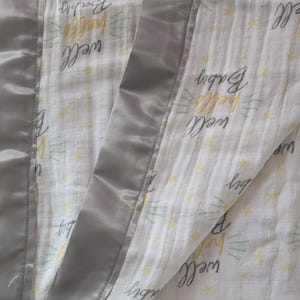 May include: A baby blanket with a white background and the words "well hello Baby" in a handwritten style. The blanket has a gray satin border and a small tag with text.