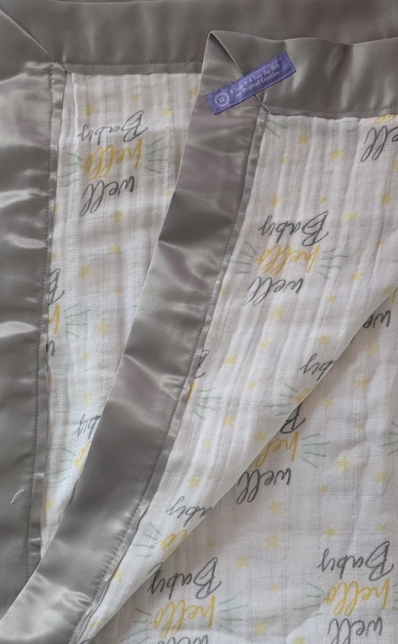 May include: A baby blanket with a white background and the words "well hello Baby" in a handwritten style. The blanket has a gray satin border and a small tag with text.