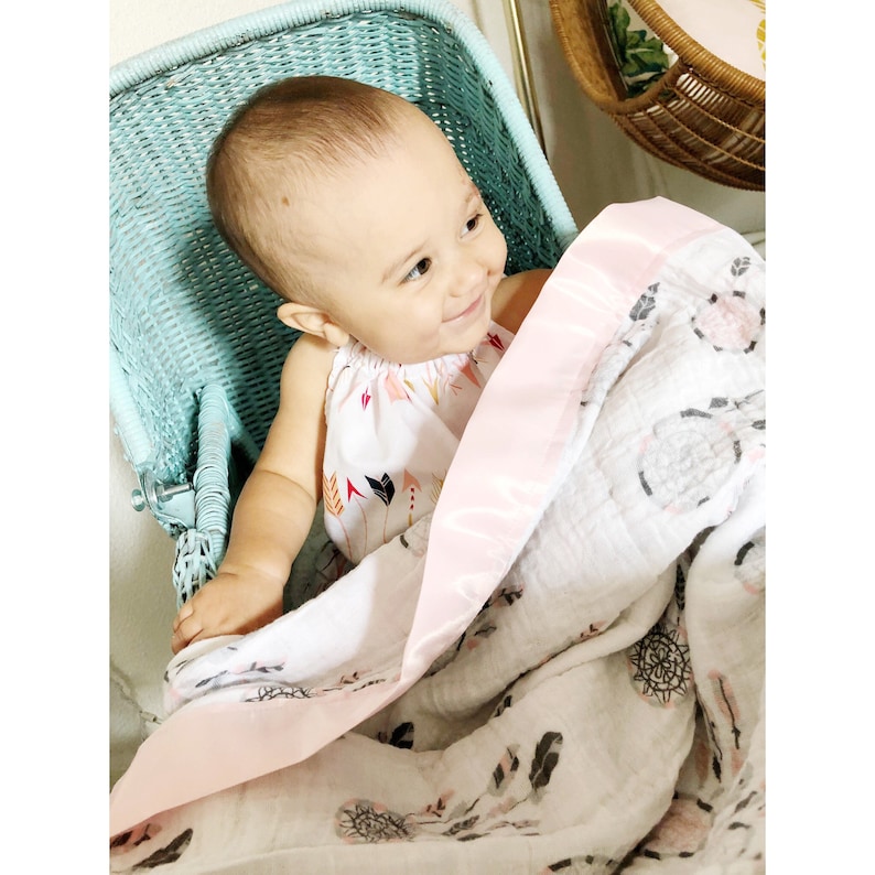 May include: A baby in a light blue wicker chair smiles, wrapped in a white and pink blanket. The blanket has a pink satin border and a pattern of feathers and dream catchers. The baby wears a white halter top with a colorful arrow design.