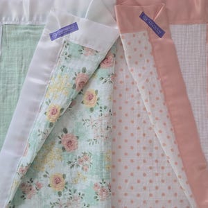 May include: Close-up of two baby blankets. One has a white satin border with a mint green interior and a floral pattern. The other has a pink satin border with a white interior and a pink polka dot pattern. Both have a purple tag.