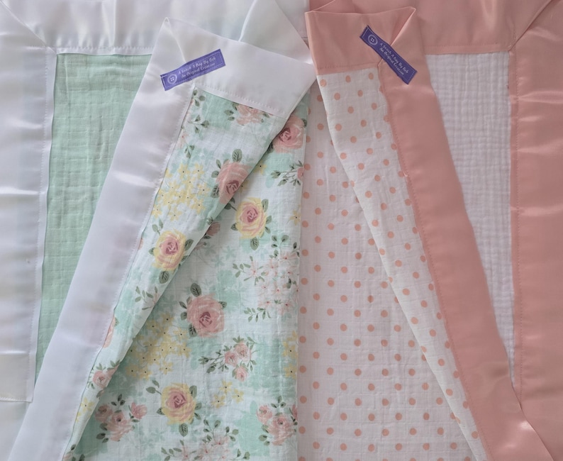 May include: Close-up of two baby blankets. One has a white satin border with a mint green interior and a floral pattern. The other has a pink satin border with a white interior and a pink polka dot pattern. Both have a purple tag.