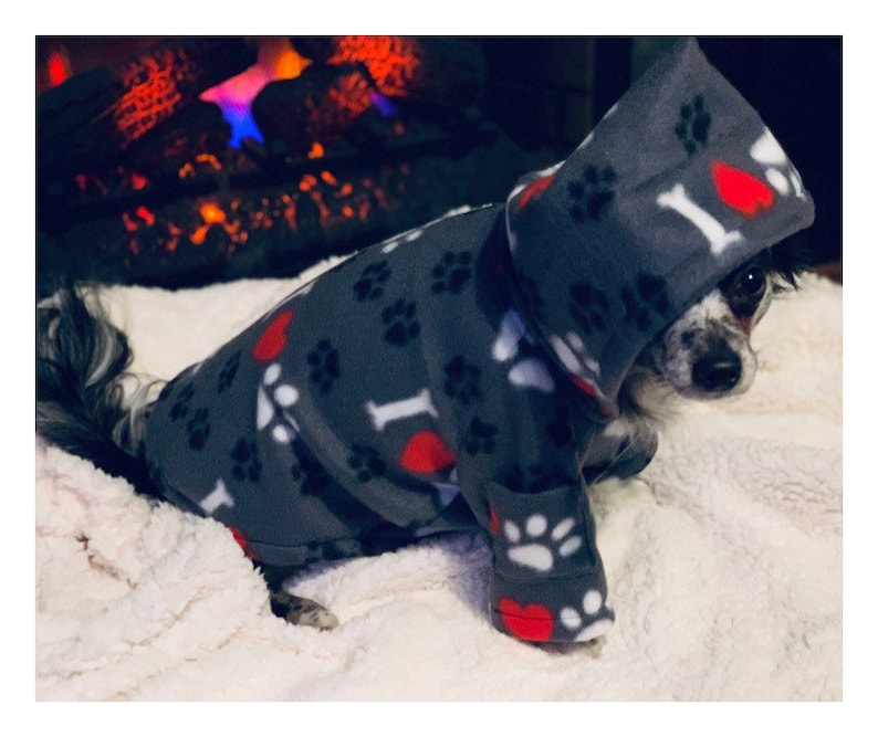 May include: A small dog wearing a gray hooded fleece sweater with a paw print and heart pattern. The sweater has the text "I love" on the front. The dog is lying on a white fluffy blanket.