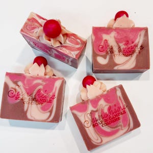 May include: Four bars of handmade soap with a swirl design in shades of pink, white, and brown. Each bar is topped with a decorative cream-colored swirl and a red cherry. The soap bars are labeled "Sunshine Soap & Candle Company."